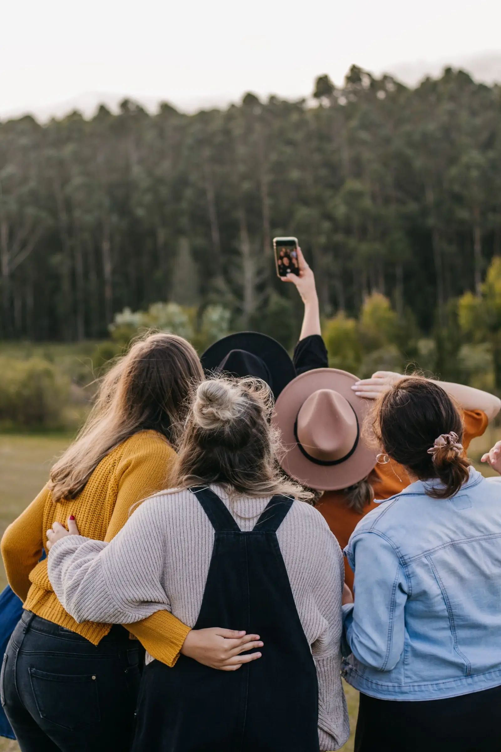 Home Back view of unrecognizable group of friends taking self portrait on smartphone while standing close to each other near green forest during trip in countryside